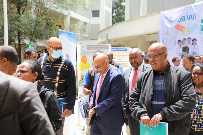 A mixed group of people walk amongst posters on display in a sunny, outdoor courtyard